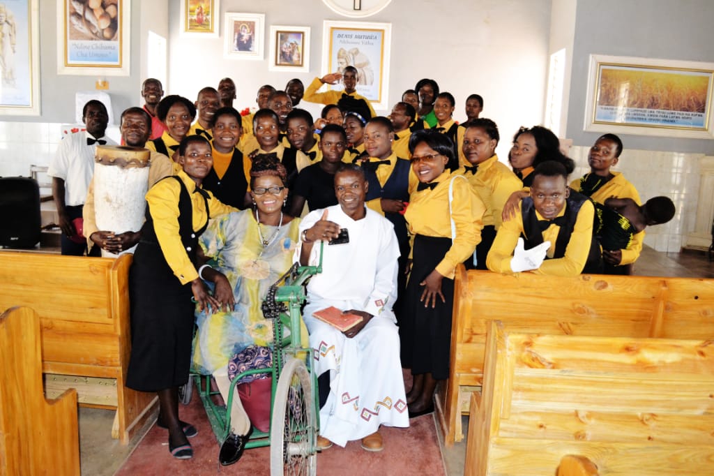 Second image of the community with lady Mayimayi in her wheelchair with the choir and the parish priest seated to her left. Everybody smiling.