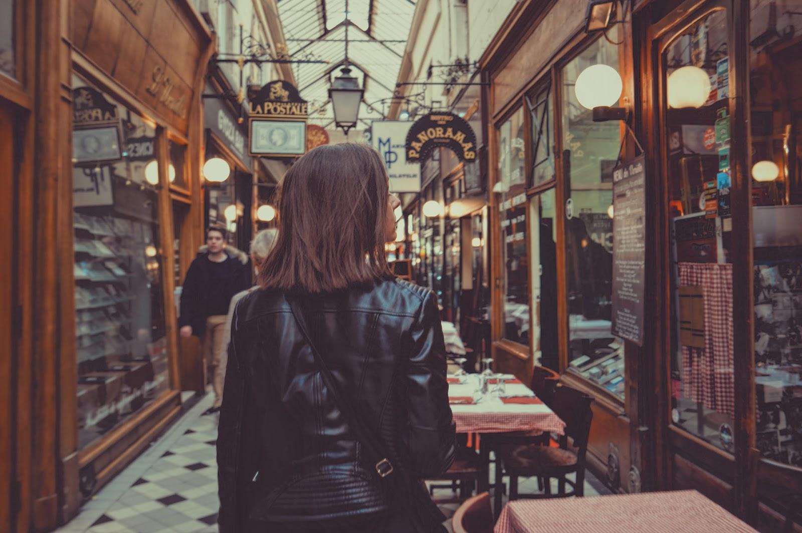 La imagen muestra a una mujer de espaldas a la imagen. Ella es blanca, tiene el cabello castaño y liso a la altura de los hombros. Esta mujer camina por un pasillo lleno de tiendas. Se pueden ver algunas mesas de un restaurante cerca de ella.