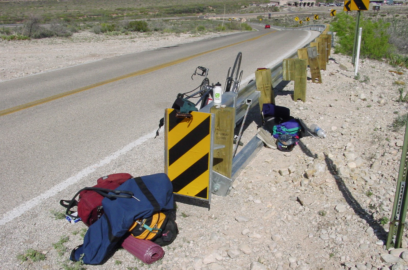 Bags strewn on the side of the road with a bicycle upside down leaning on a guardrail. Desert surrounds. 