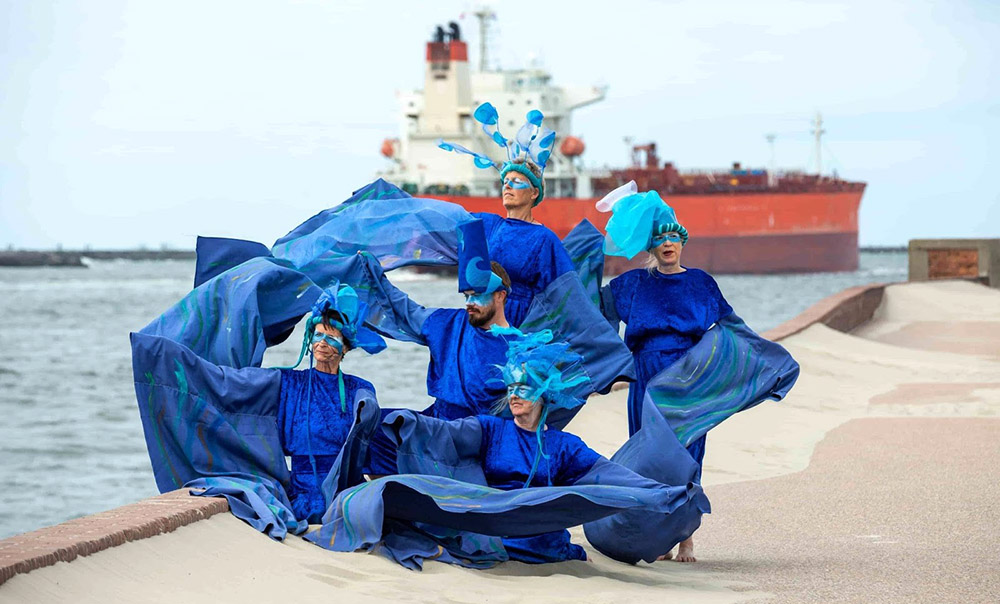 Ocean Rebels in blue robes pose by a rive with a tanker behind