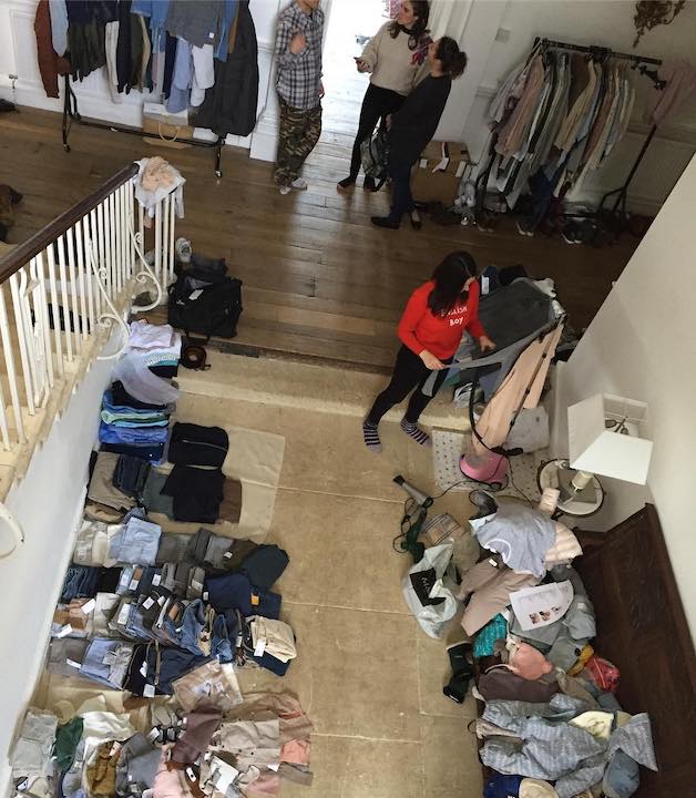 Behind the scenes image of a photoshoot in a house where one room is filled with clothing. There is a wardrobe stylist steaming some items hanging on a rack.