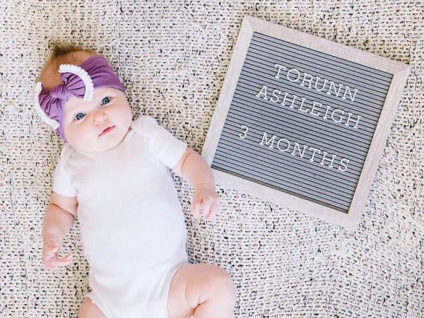 a gray letterboard with text "torunn ashleigh 2 months" and baby wearing purple headband lying to the side