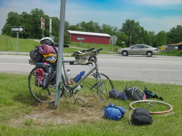 Bicycle loaded for camping with its front wheel removed and the front bags strewn along the side of a road. 