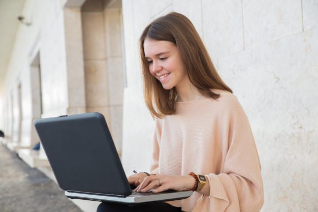 Smiling teen girl browsing on laptop on bench at building Free Photo