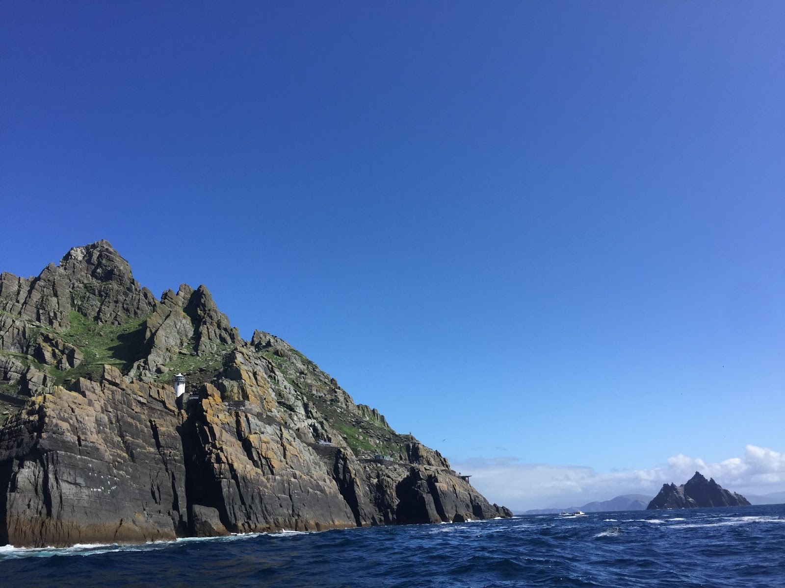 Photo of the Skellig Islands taken from the boat; one is near and the other in the distance. The rocks rise sharply out of the deep blue water. 