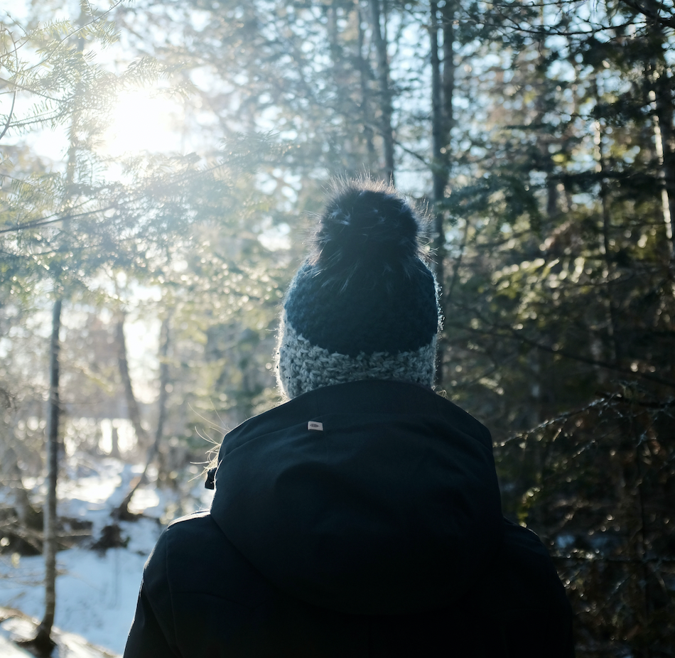 Back profile of a person with a dark winter coat and hat, sunlight peaking through the trees, in a wintery forest