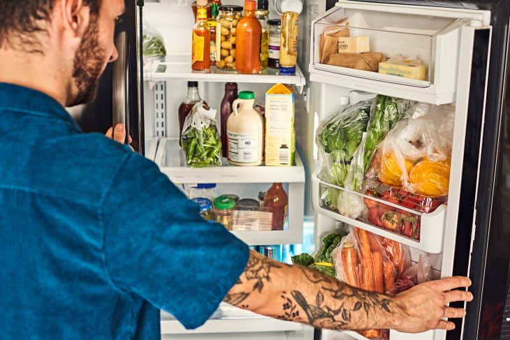 A man standing in front of an open two-door frisge. Over his shoulder one side of the fridge is visible. The door shelves are full of bagged produce, and the crisper drawer that is visible contains a variety of condiment jars. 