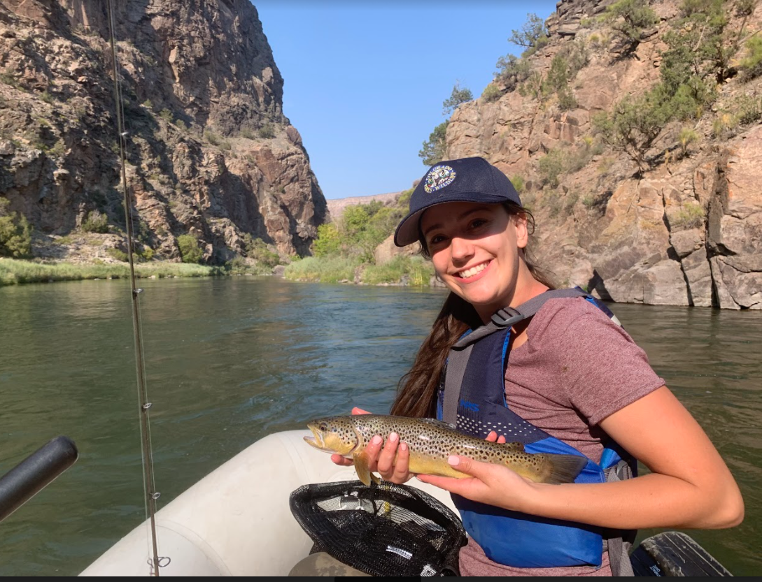 A young woman with a navy Colorado Parks and Wildlife hat holds up a fish and smiles as she sits in a raft on a river.