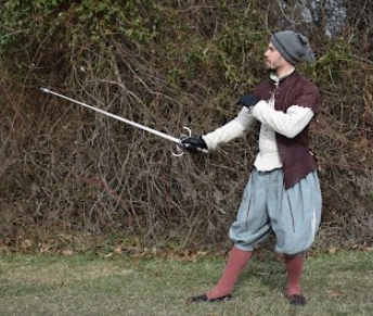 A man wearing a burgundy doublet, blue pants, red socks, and grey hat stands in profile with his right leg outstretched, holding a sword in front of him, with a backdrop of bare wooden vines
