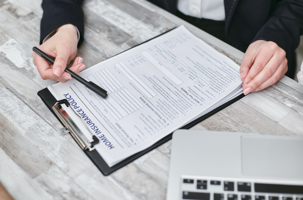 A person's arm is resting on a white wooden table. There is a black pen in their hand, and they are pointing the pen to a clipboard with an insurance policy form. The corner of a laptop keyboard is visible next to the clipboard.