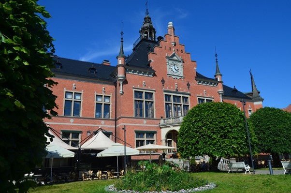 A prestigious building in the center of Umea with a garden in the foreground and the entrance to the pink building with a dark roof in the background. A large tree on the left.