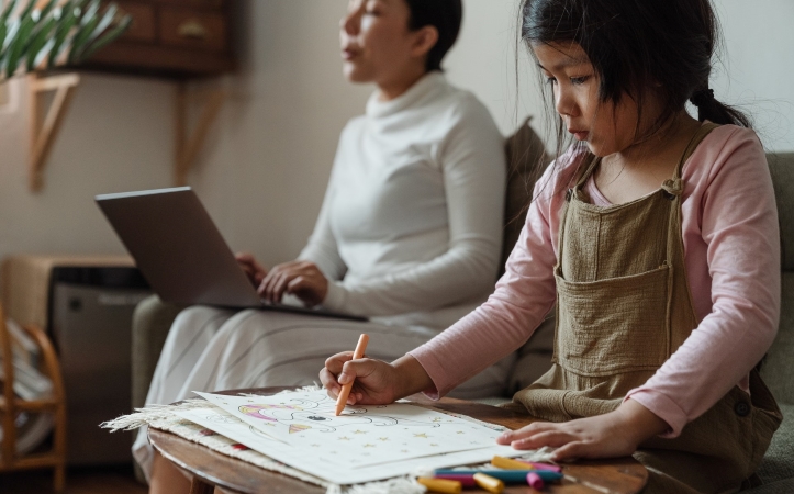 A mother working from home while her young daughter colors nearby.