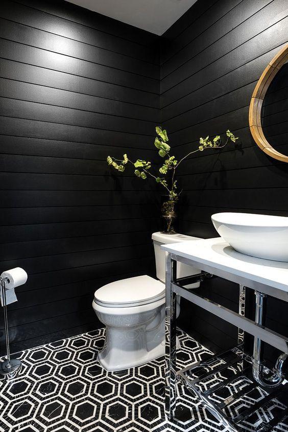 Banheiro com revestimento de madeira preta nas paredes, piso geométrico preto e branco, louças brancas, espelho com moldura de madeira e acessórios pratas.