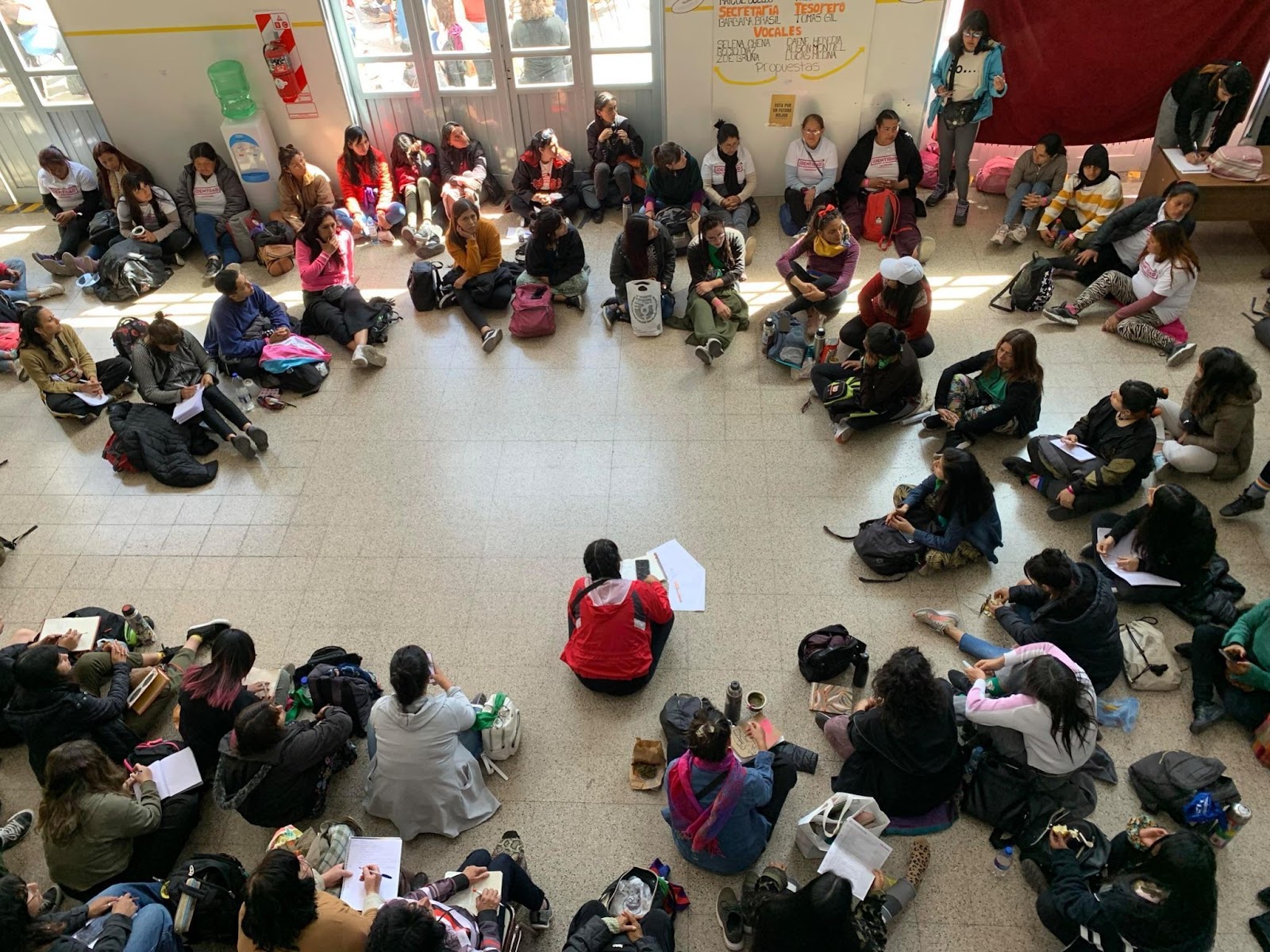 Bird’s eye view of a large group of people sitting in a circle on a warm gray ceramic tiled floor.