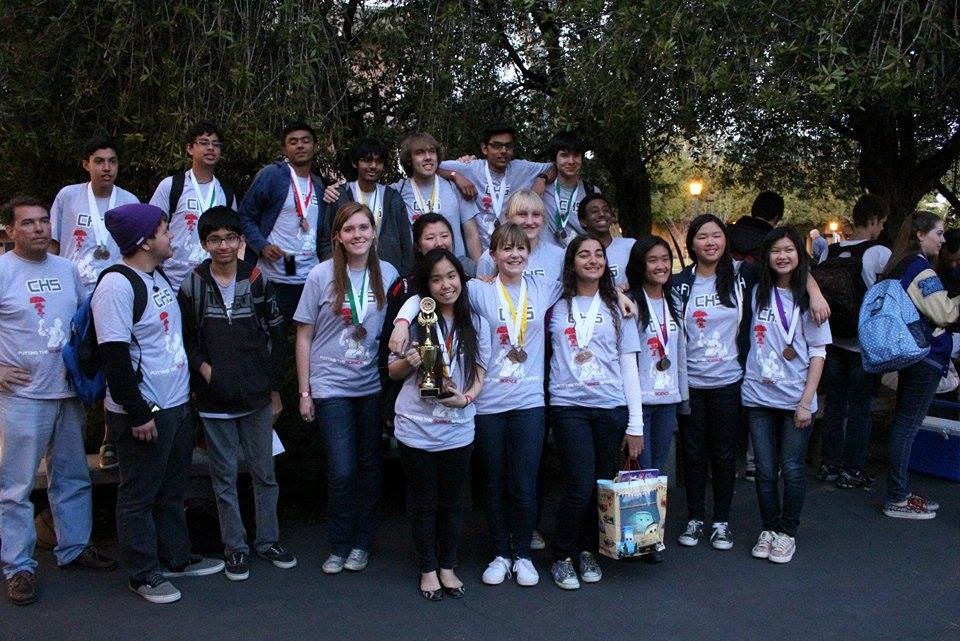 A group of high schoolers in matching shirts with medals and a trophy, posing for a group photo.