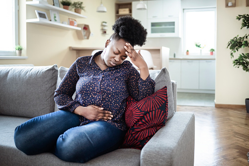 A black woman sitting on her living room couch, one hand holding her stomach due to cramps and head resting on her other hand as her eyes are closed. This image represents feeling distressed, anxious, and overwhelmed. You can get help from an anxiety therapist in Los Angeles, CA for anxiety treatment to help with overcoming obsessive-compulsive disorder in Los Angeles, CA too. 91364