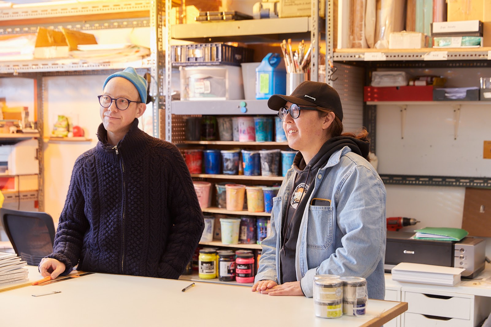 Image: Nick Butcher, left, and Nadine Nakanishi, right, in Sonnenzimmer's studio. They stand at a mostly bare beige table. Behind them are metal racks with multi-colored pint tubs of ink and other tools. To the right, a printer with a green folder on it and a drill. Photo by Sarah Joyce.