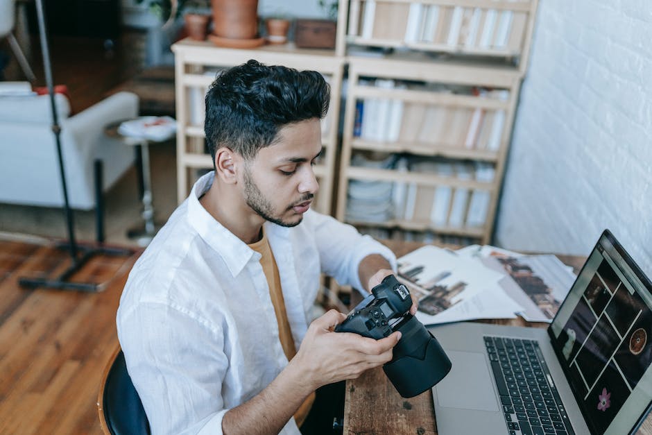 Side view of Indian male photographer in casual clothes watching photos on photo camera while sitting at table and browsing laptop at home in daytime