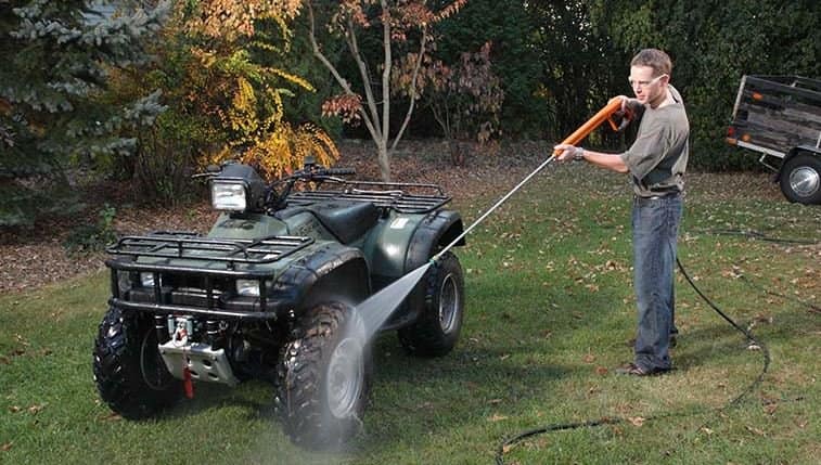 A man in his lawn giving his ATV a wash, by using a powerwasher. This is to help upkeep maintenance with the ATV.