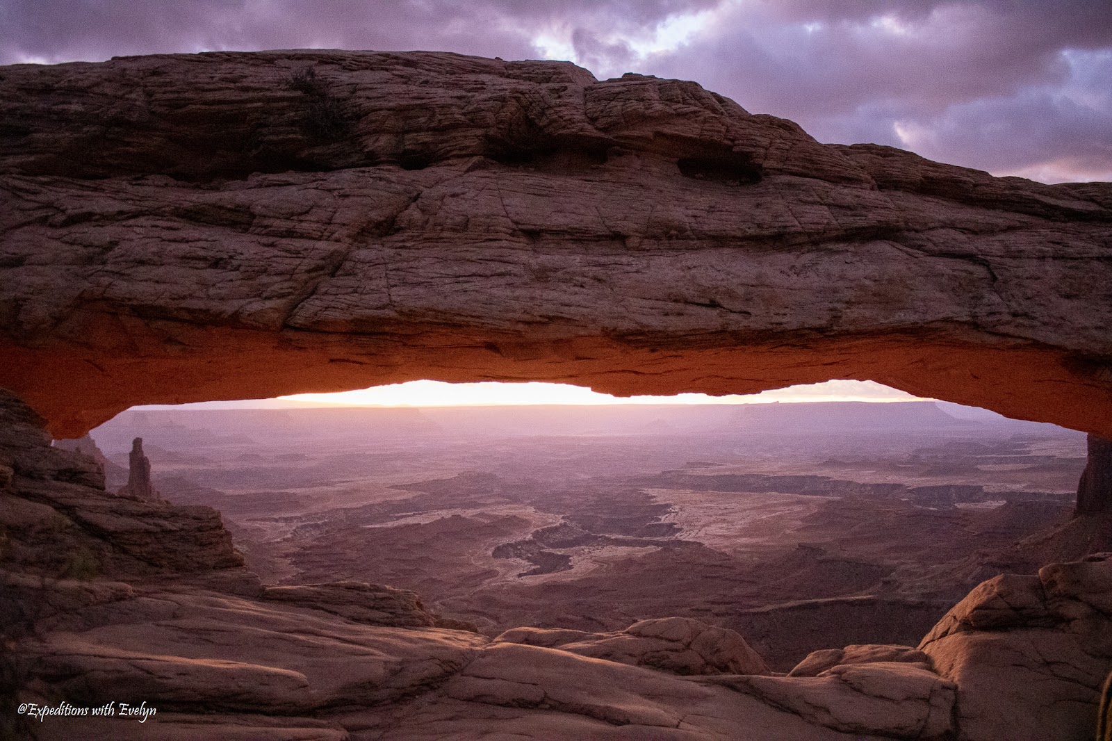 Pink and purple hues of sunrise fill a canyon behind Mesa Arch.  Observing this scene is on of the most popular things to do in Moab Utah.