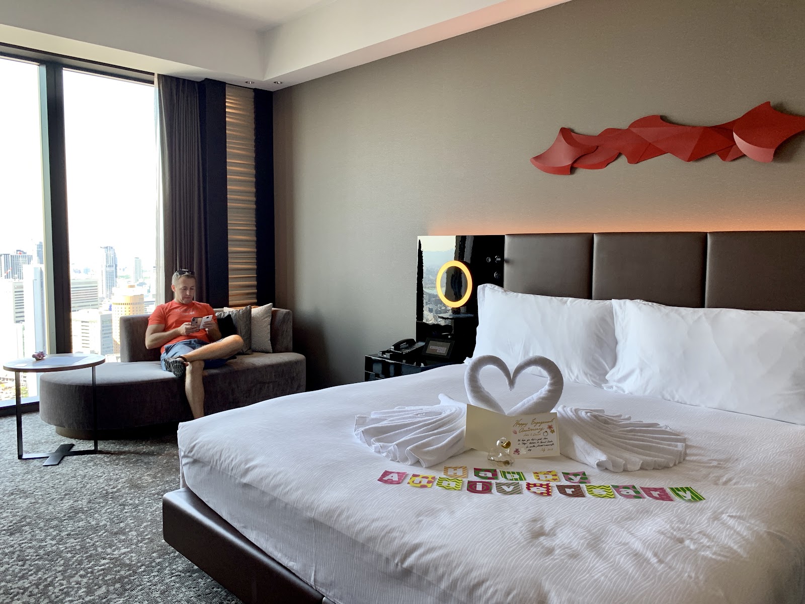 hotel room with "Happy Anniversary" displayed on bed, and man sitting on couch