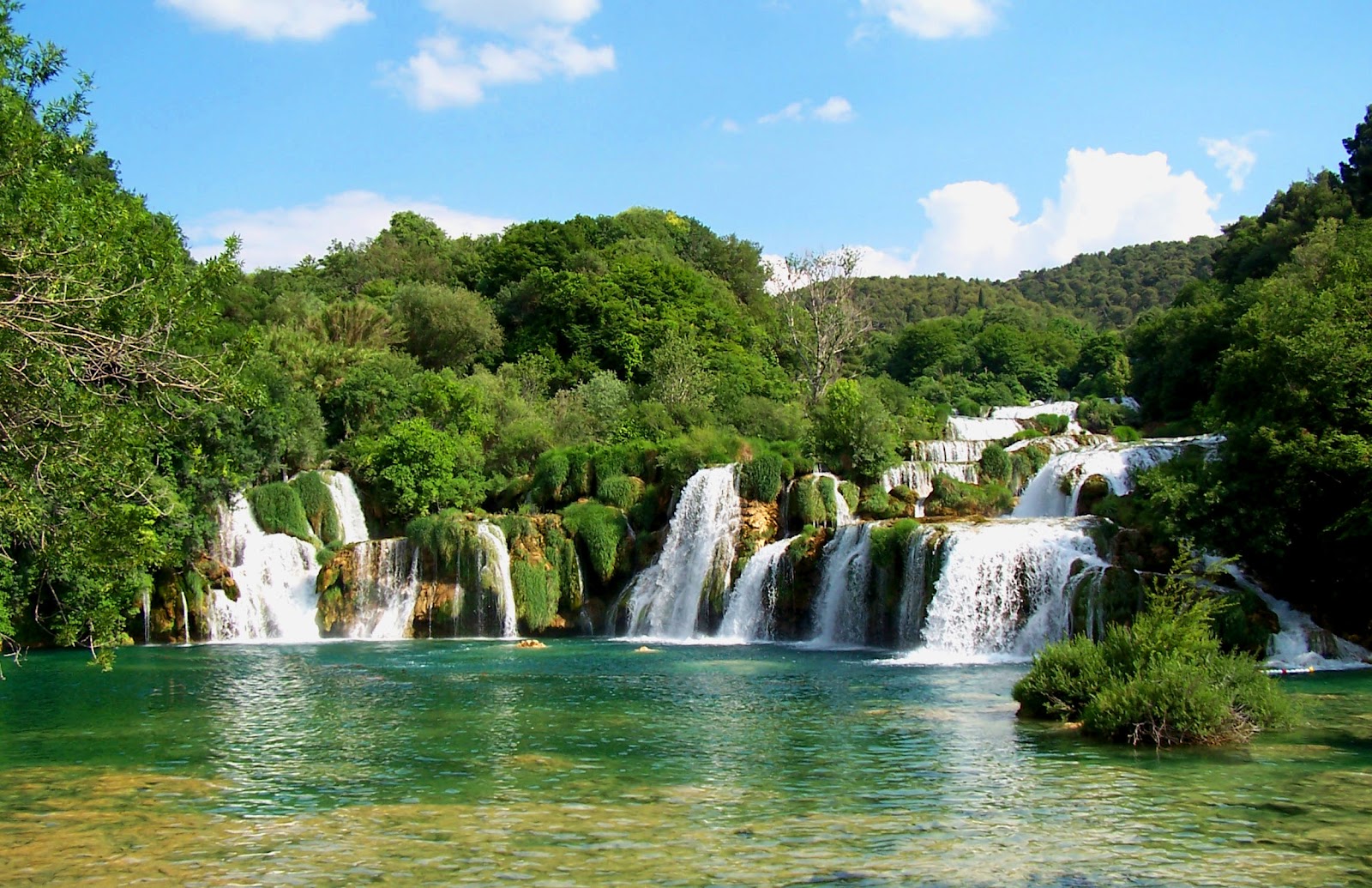 wide waterfall white foamy water going into crystal clear, secluded emerald lake, surrounded by trees. Sunny day in krka national park croatia