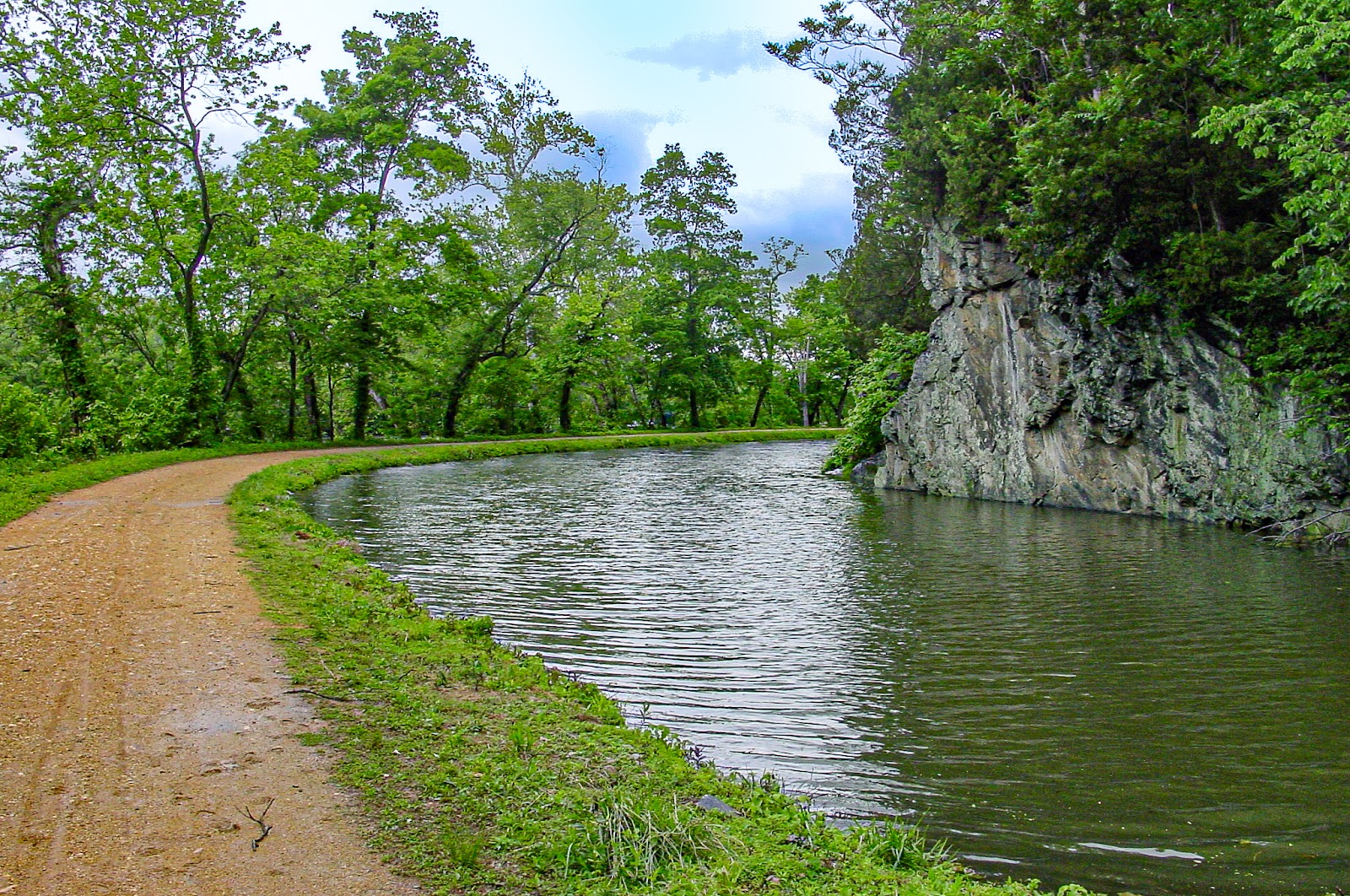 A brown road passes through green grass beside a placid waterway with rock cliff opposite the road. 