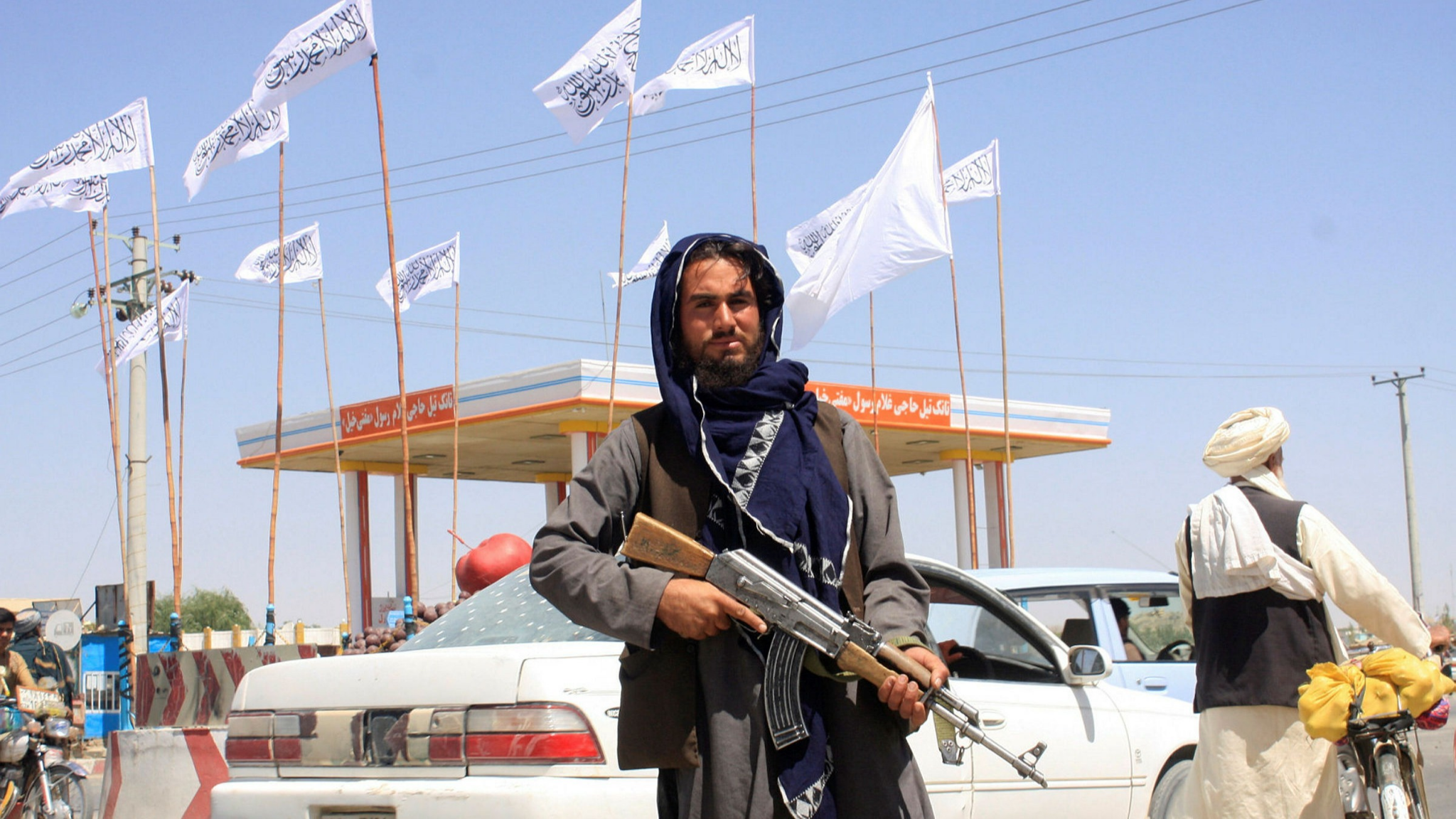 A man in hooded Middle-East clothing stands holding a rifle in front of a background of white flags with symbols, blowing in a light wind at a gas station.