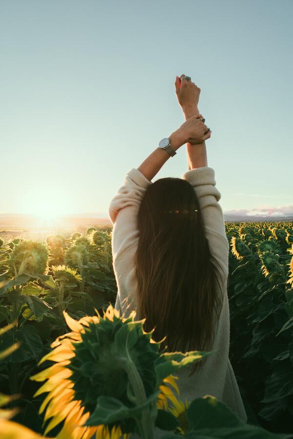 Foto de uma mulher em um campo de girassóis, de costas, com as mãos para cima. 