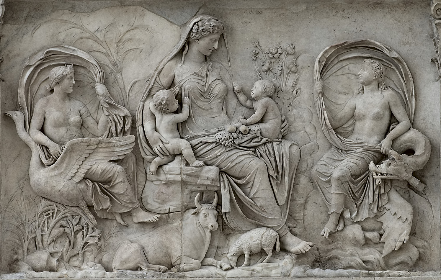 The Augustan Ara Pacis, 13 BCE, is an exemplary representation of the state Greco-Roman style, featuring a symbolic scene with three women. The central figure holds two infants in her lap, and at their feet are animals. 