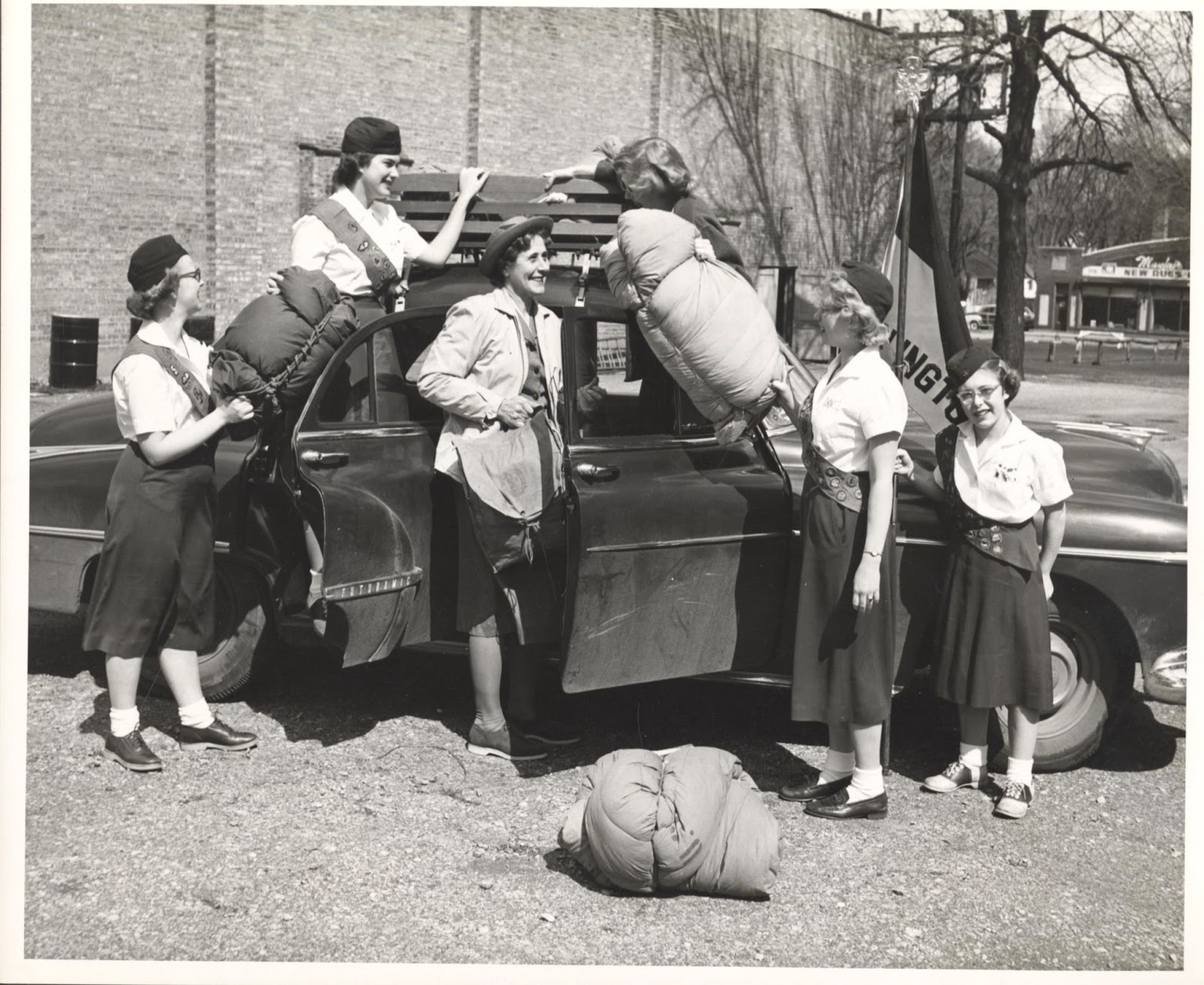 Six people loading up a car with sleeping bags, two of the people are standing on the car's doorframe with the doors open and the other three are standing on the ground. Five of them are wearing girl scout uniforms and the person in the middle, Opal Weimer, is wearing a hat and coat and holding a bag at the door of the car.