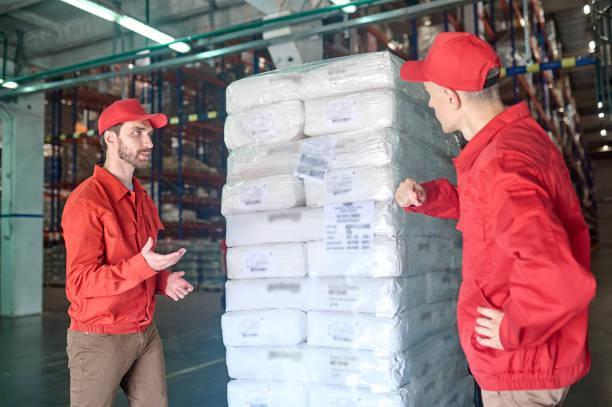 Two storehouse workers talking about the transportation of goods Serious young warehouse employee having a conversation about the cargo delivery with his male colleague Optimizing Supply Chains: The Role Of Cold Storage Services stock pictures, royalty-free photos & images