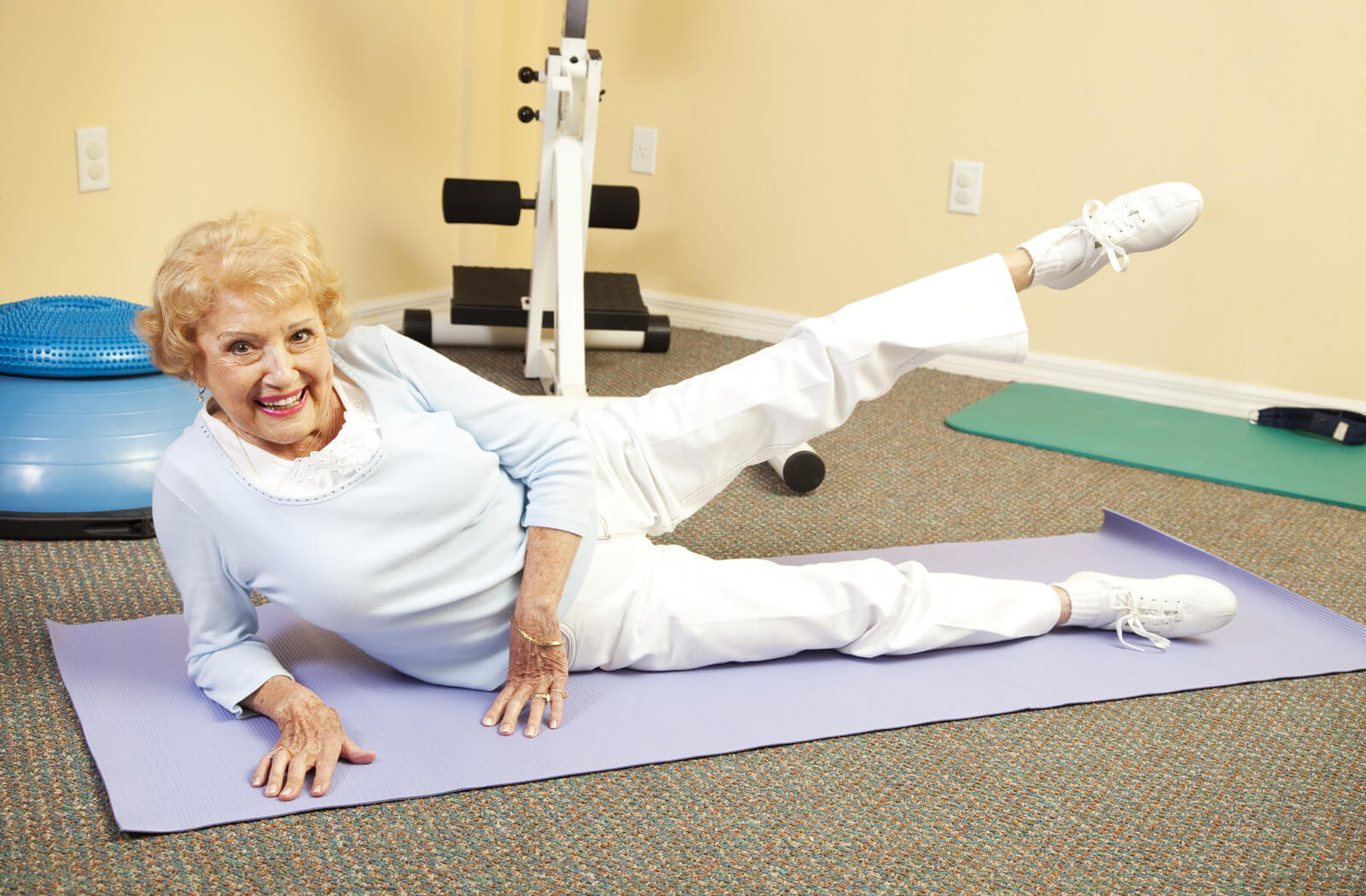 a senior woman lying on her side on a yoga mat performs a prone leg raise with her left leg while smiling at the camera