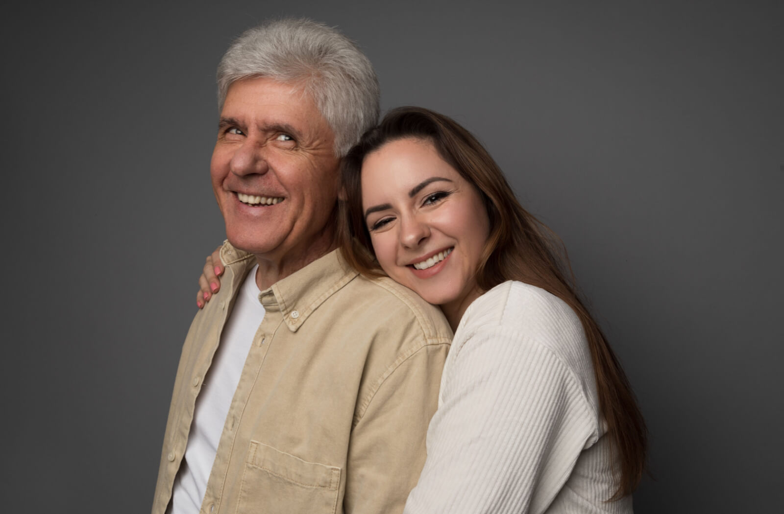 A senior man grinning as his daughter embraces his shoulder with a warm smile.