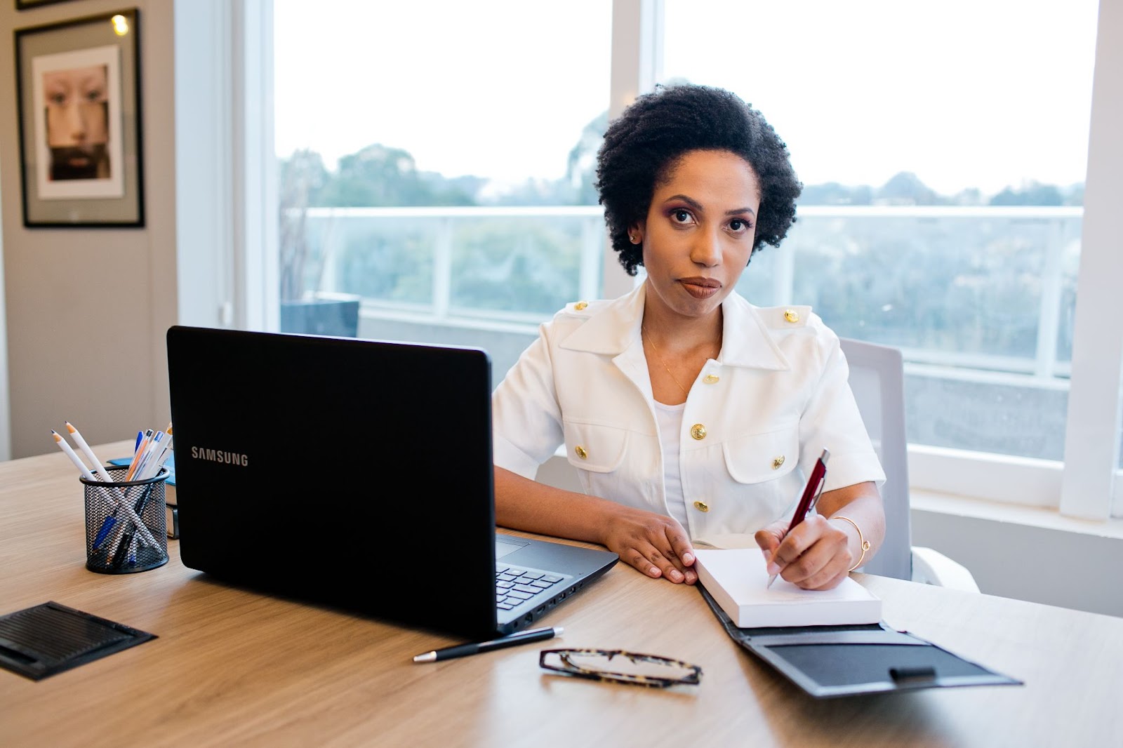 Imagem mostra Anna Lyvia Ribeiro, uma mulher negra em uma sala de escritório. Ela está sentada e segura uma caneta na mão e seu notebook a sua frente. 