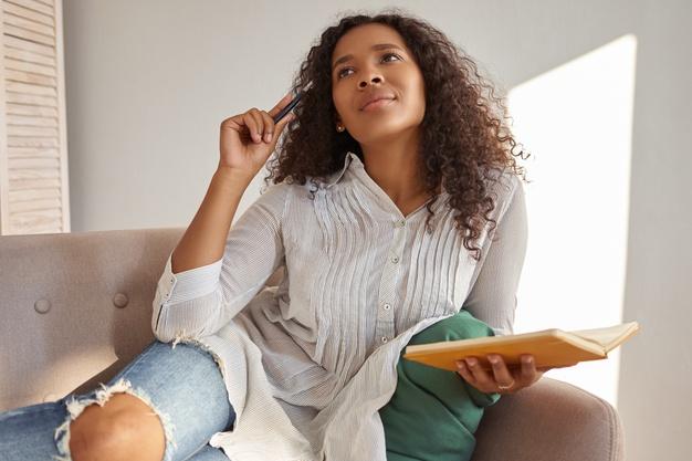 Indoor shot of beautiful young afro american lady in blouse and ripped jeans having pensive facial expression, looking up, scratching head with pen, making notes in copybook, developing business plan Free Photo
