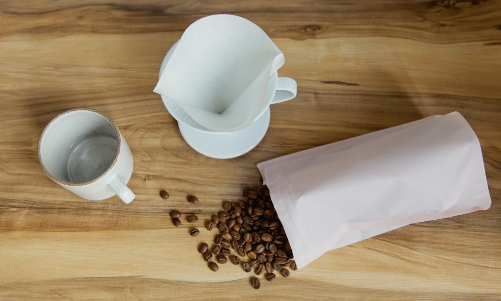 White V60 with filter paper sits on wooden counter beside white mug and white multilayer coffee pouch with dark roasted coffee beans on table. 