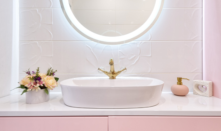 A pink and white sink and vanity are illuminated by a circle mirror with a light that surrounds it. The vanity has matching pink and gold decor, as well as a small bouquet of various pink flowers. 