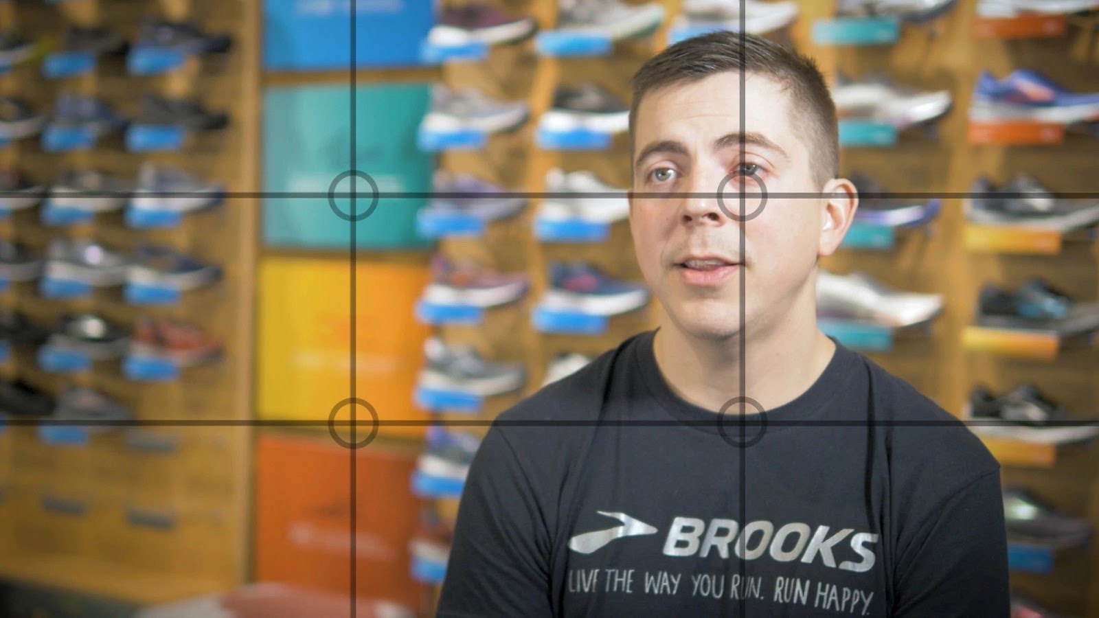 Still video frame of a customer case study interview with lines on the screen depicting the rule of thirds. The still is of a man in a black Brooks Running t-shirt with a wall of shoes blurred out in the background. 
