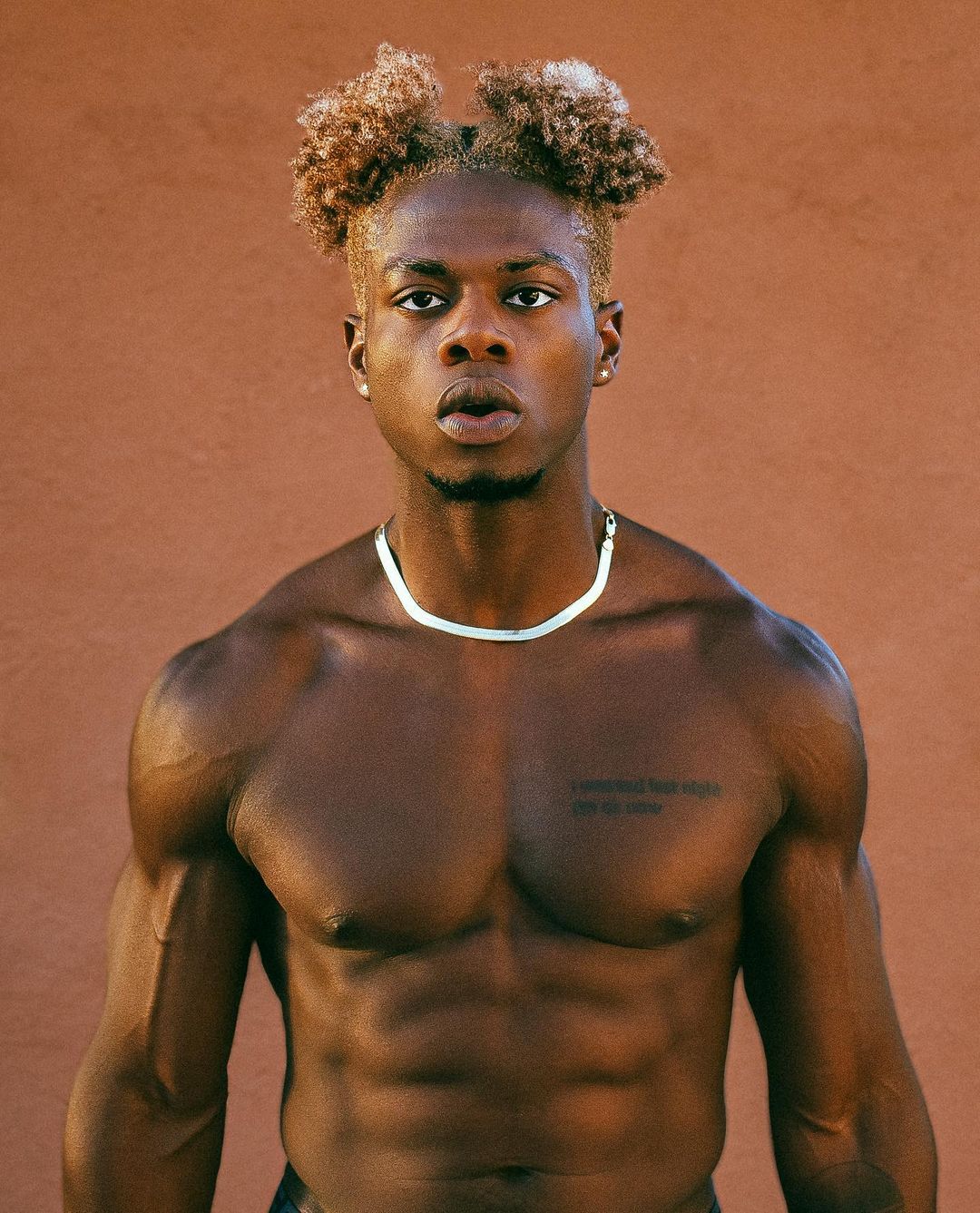 Curly haired man posting with no shirt while wearing a gold chain