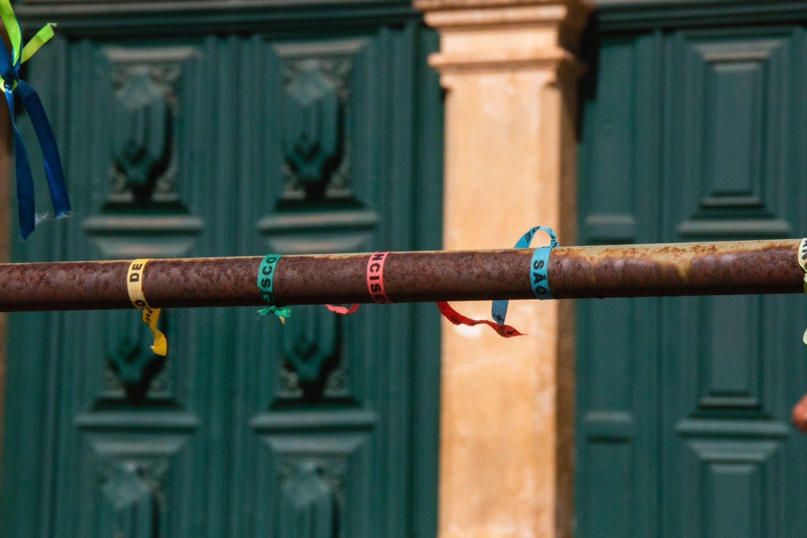 Barra de ferro com quatro pulseiras coloridas, típicas do nordeste brasileiro. Ao fundo, duas grandes portas coloniais em tom de verde escuro.