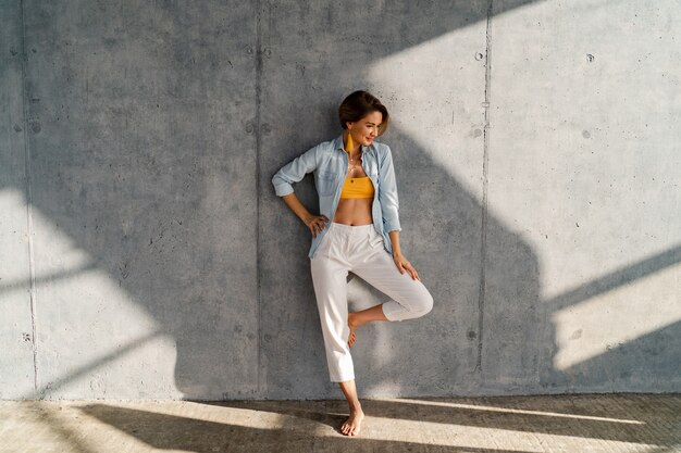 A woman standing confidently in front of a sunlit concrete wall. She is dressed in a denim shirt, a vibrant yellow top, and white bottoms