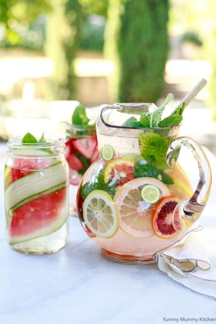 Three detox water recipes in glass pitchers sit on an outdoor table. A glass pitcher with citrus infused detox water, a jar with cucumber and watermelon, and another jar with strawberries and mint.