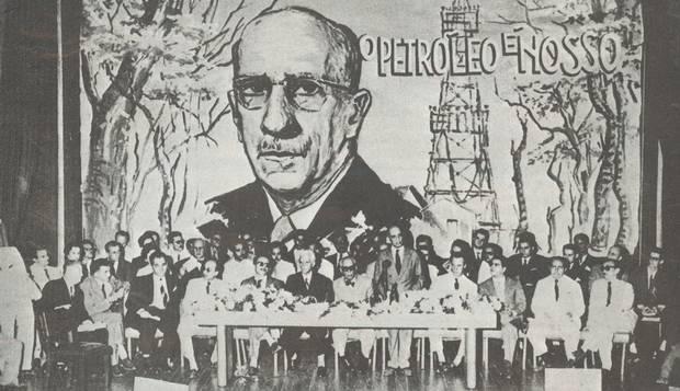 Imagem antiga em preto e branco de uma reunião de executivos com o backdrop da campanha “O petróleo é nosso”.