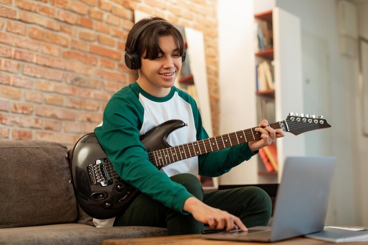 Jovem segura uma guitarra enquanto mexe no computador, sentado em um sofá e usando fones de ouvido: há muitas formas de aprender online.