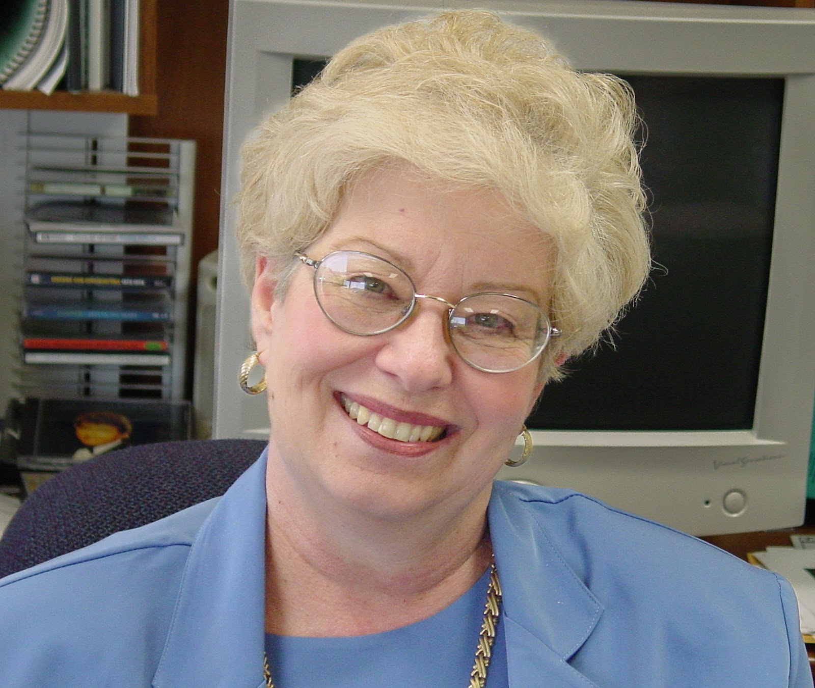 Woman in a blue dress with white hair and glasses, she smiles at the camera. 