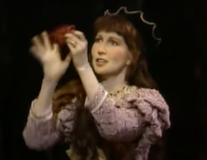 A waist-up view of a white woman with brown hair. She is dressed in a fairy tale-era costume and appears to be singing. She is crumpling a red piece of fabric in her hands.