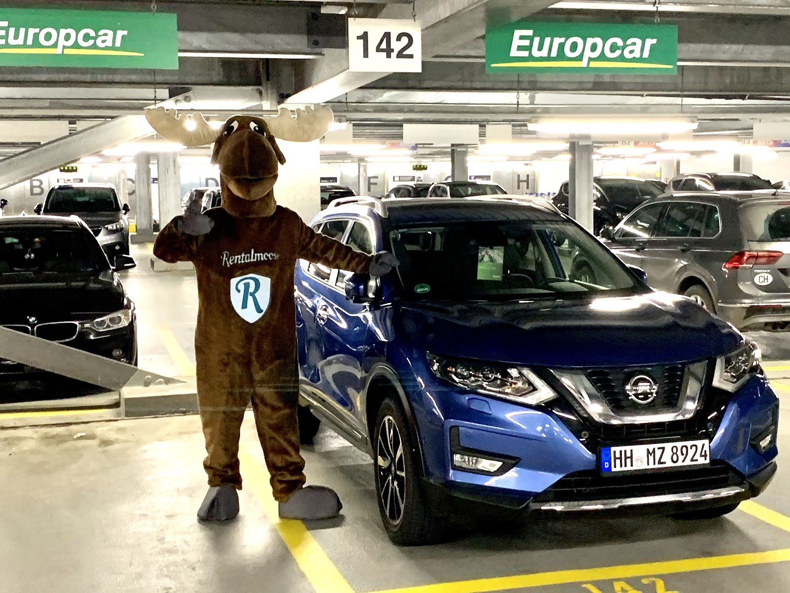 Rental Moose mascot posing in undeground garage, next to blue Nissan rental car by Europcar. Rental Moose is a global car rental broker which will help you get the best deal on your next rental car.