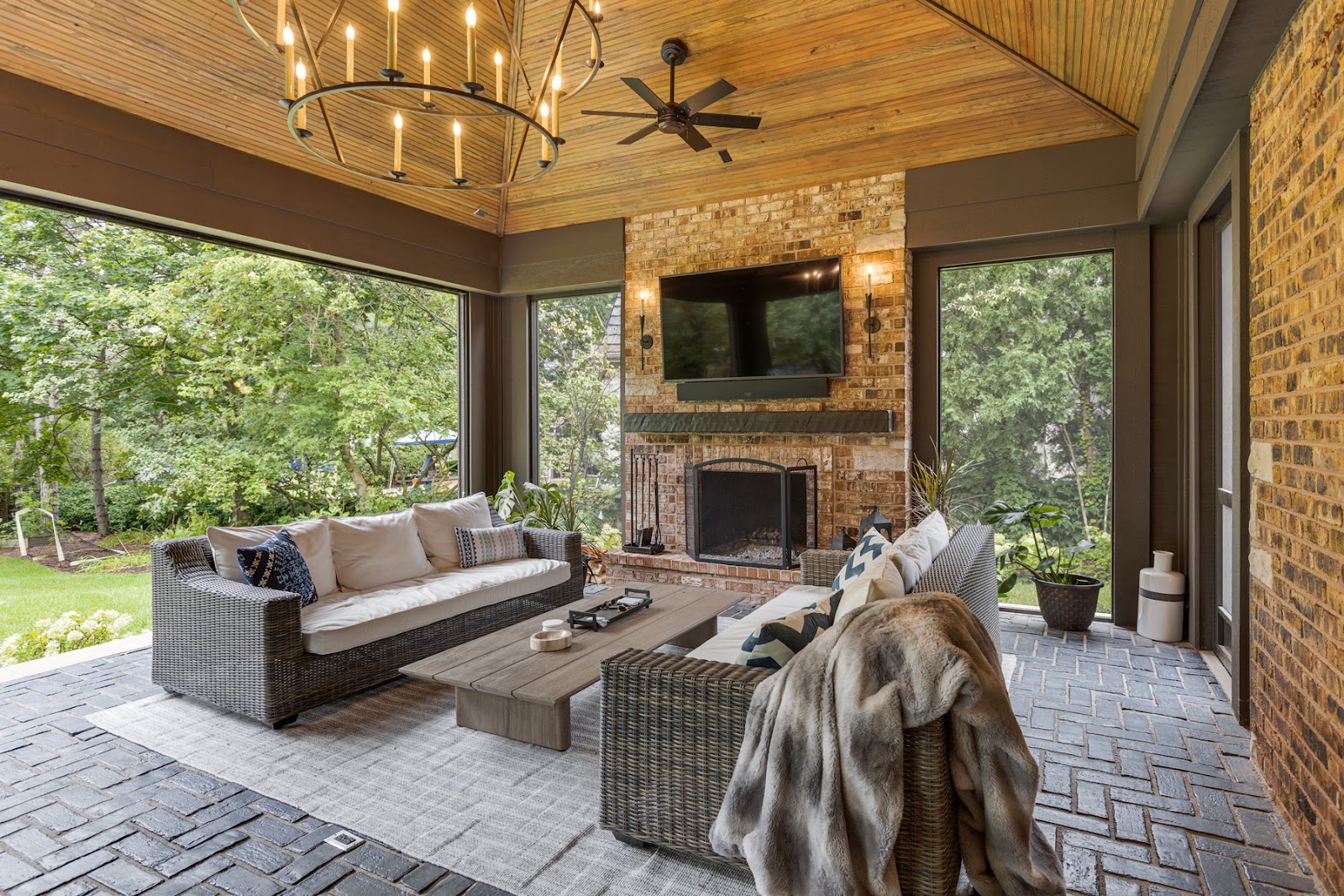 Three season room with outdoor views, a brick fireplace, wood paneled ceiling and paver flooring.