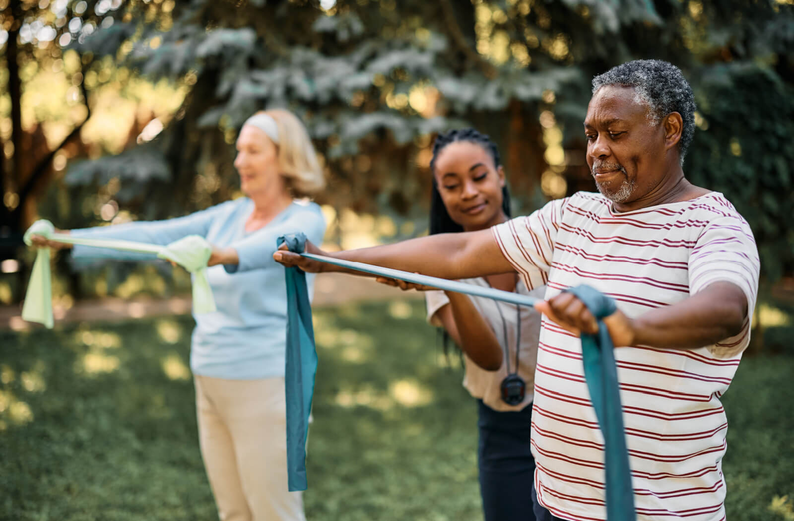A senior man and a senior woman using resistance bands during physical therapy while being assisted by a therapist in the backyard of a senior living community.